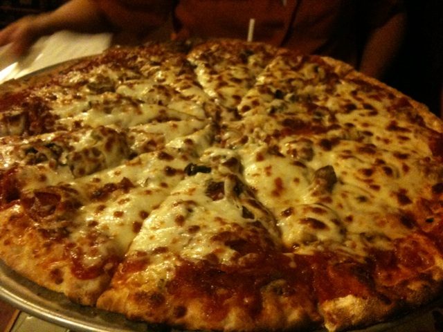 Cheese-covered pie with scattered toppings in dim light, The Works