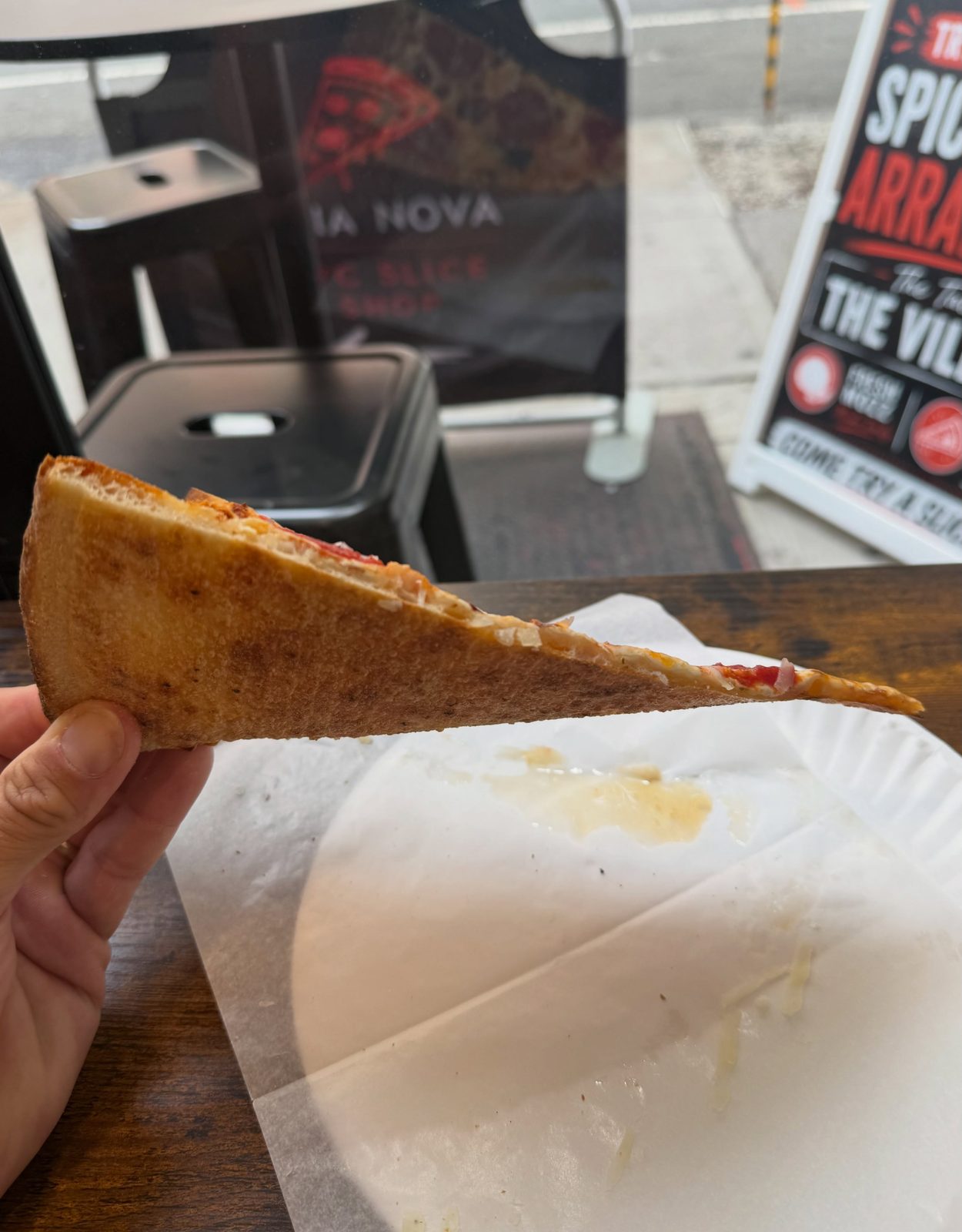 Slice held up at the window of Aria Nova Pizzeria, showing its browned underside in profile