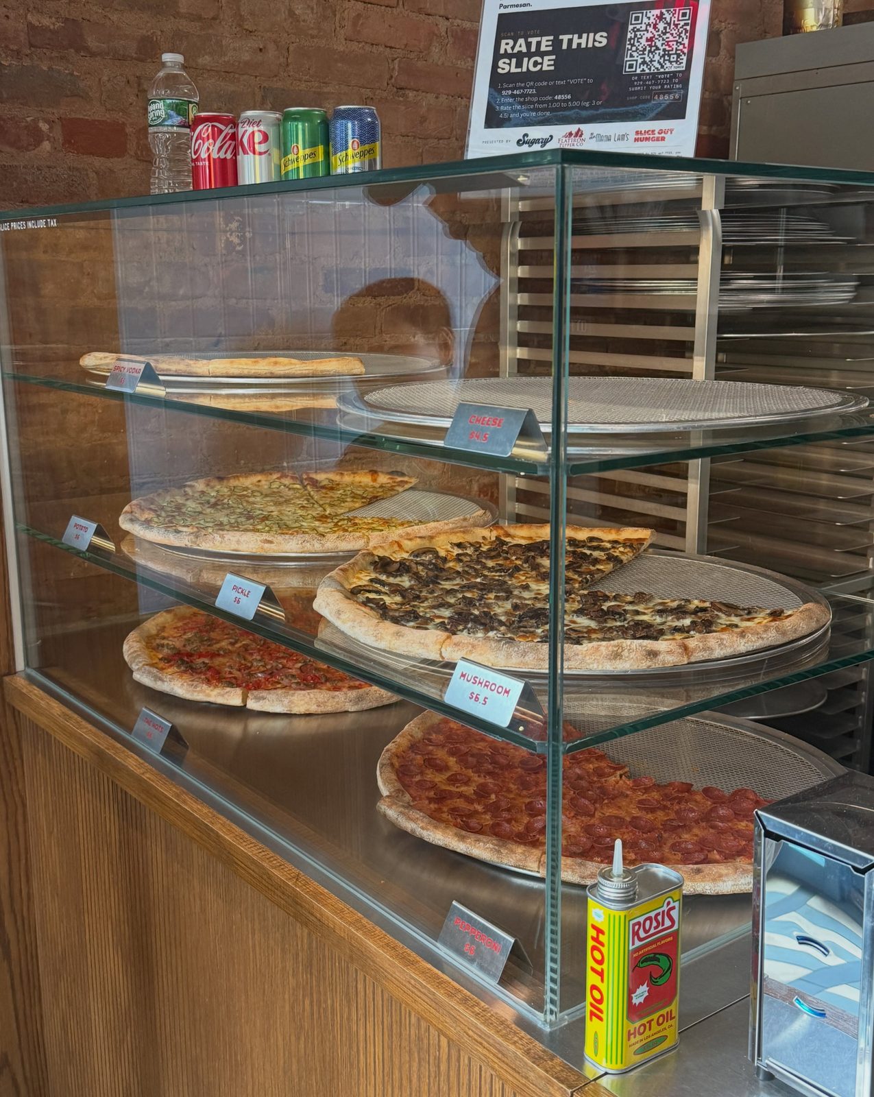 Slices in the warming case at Hots Pizza, priced by the slice under a Rate This Slice sign