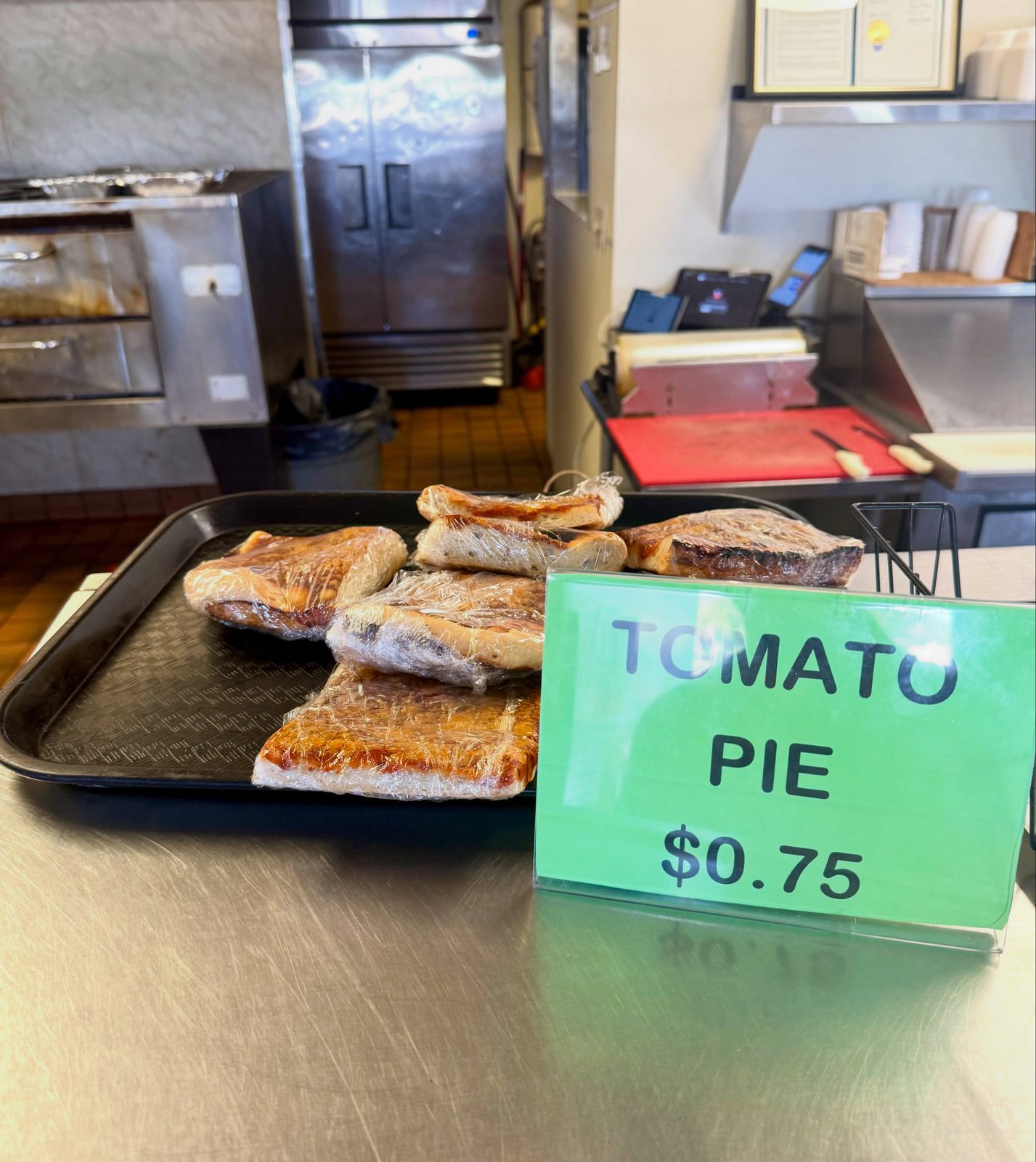 Wrapped squares of tomato pie on a tray beside a sign reading Tomato Pie 75 cents at O’Scugnizzo Pizzeria