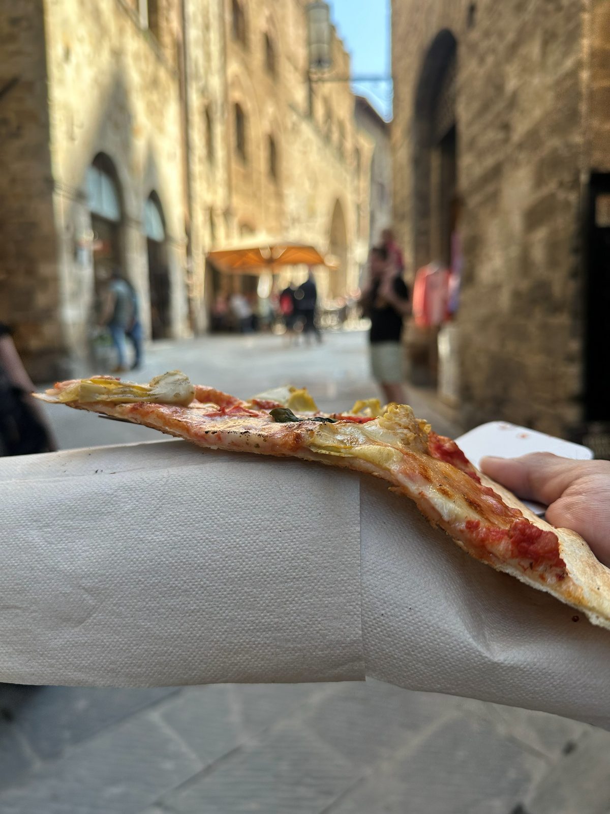 Slice held up on a San Gimignano street lined with stone towers, Ricca Pizza