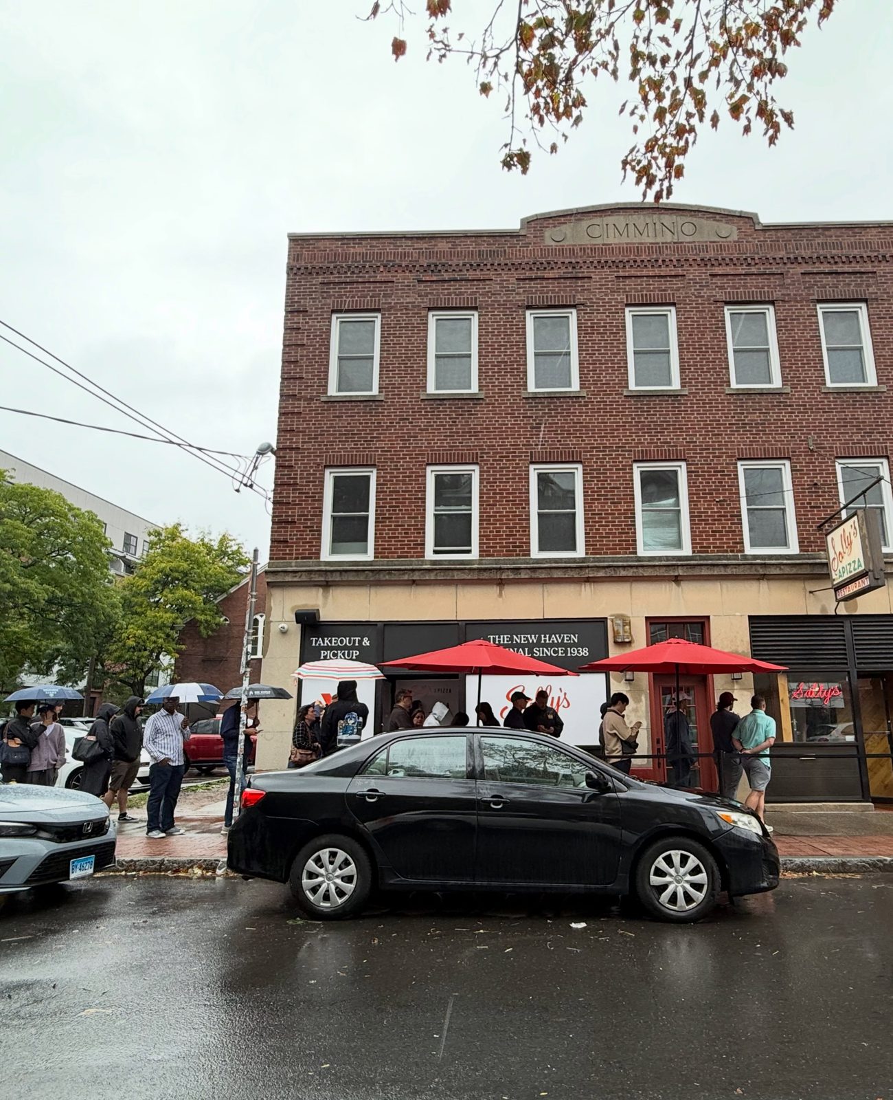 People queueing in the rain outside Sally’s Apizza on Wooster Street, below a sign reading The New Haven Original Since 1938