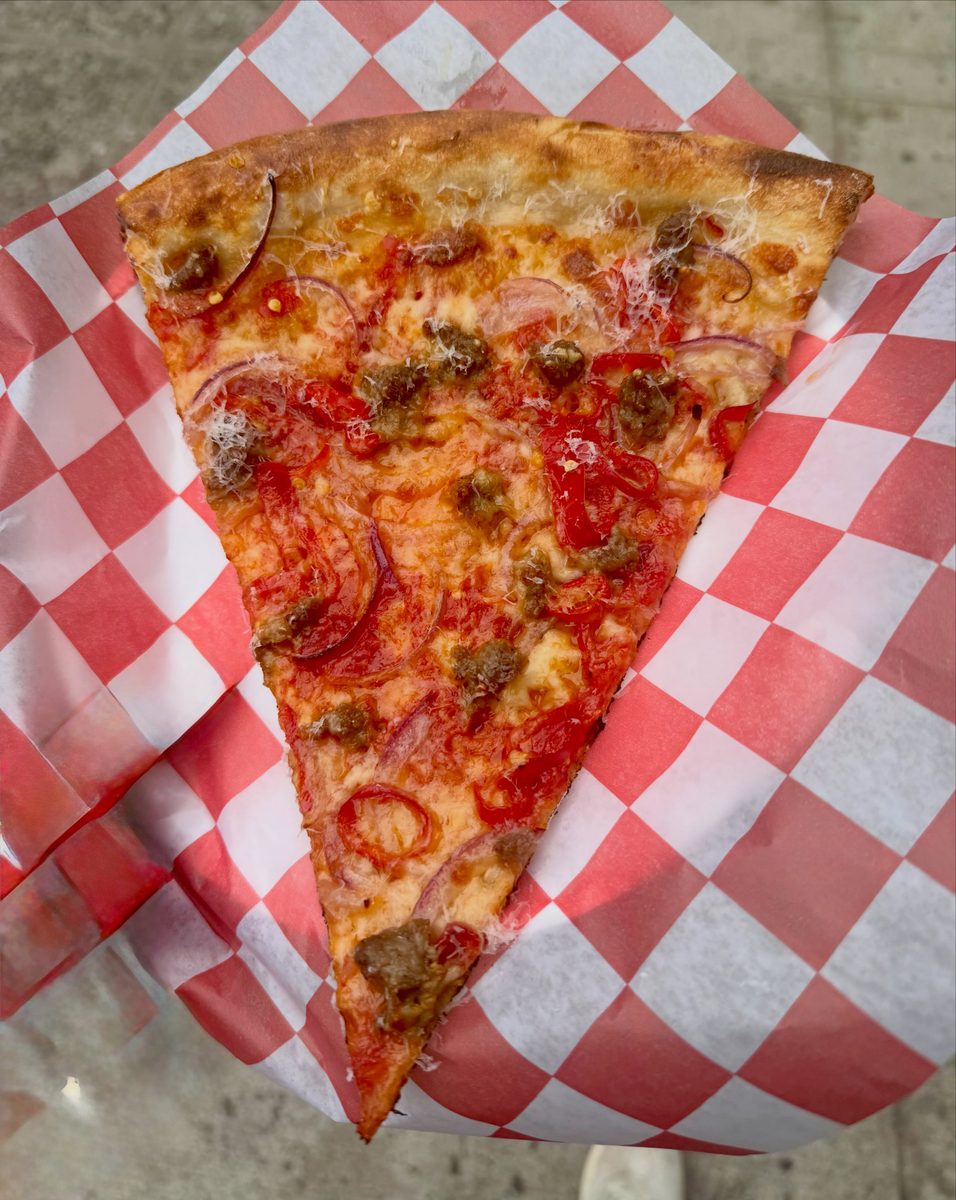 The Hots slice at Hots Pizza on checked paper, spicy sausage, hot peppers and red onion under grated cheese