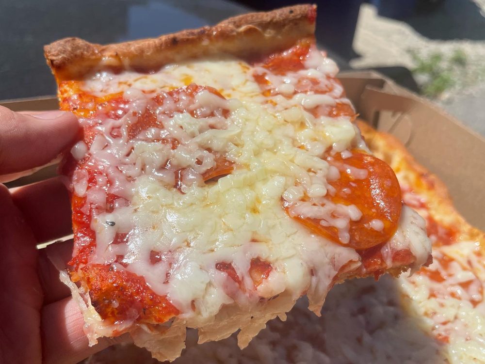 Square slice with melted cheese held in bright sunlight, DiCarlo’s Original Pizza