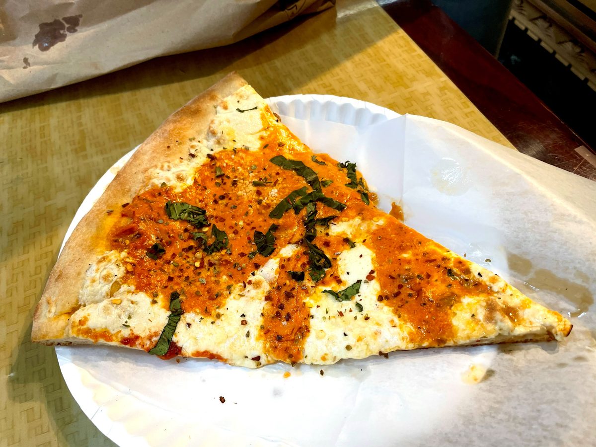 Wide New York slice on a paper plate, sauce swirled over fresh mozzarella with torn basil and chili flakes, Previti
